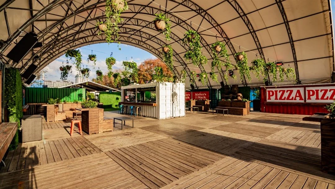 A large arched outdoor venue with a wooden deck, hanging plants, shipping container bars and a red Pizza sign on the right.